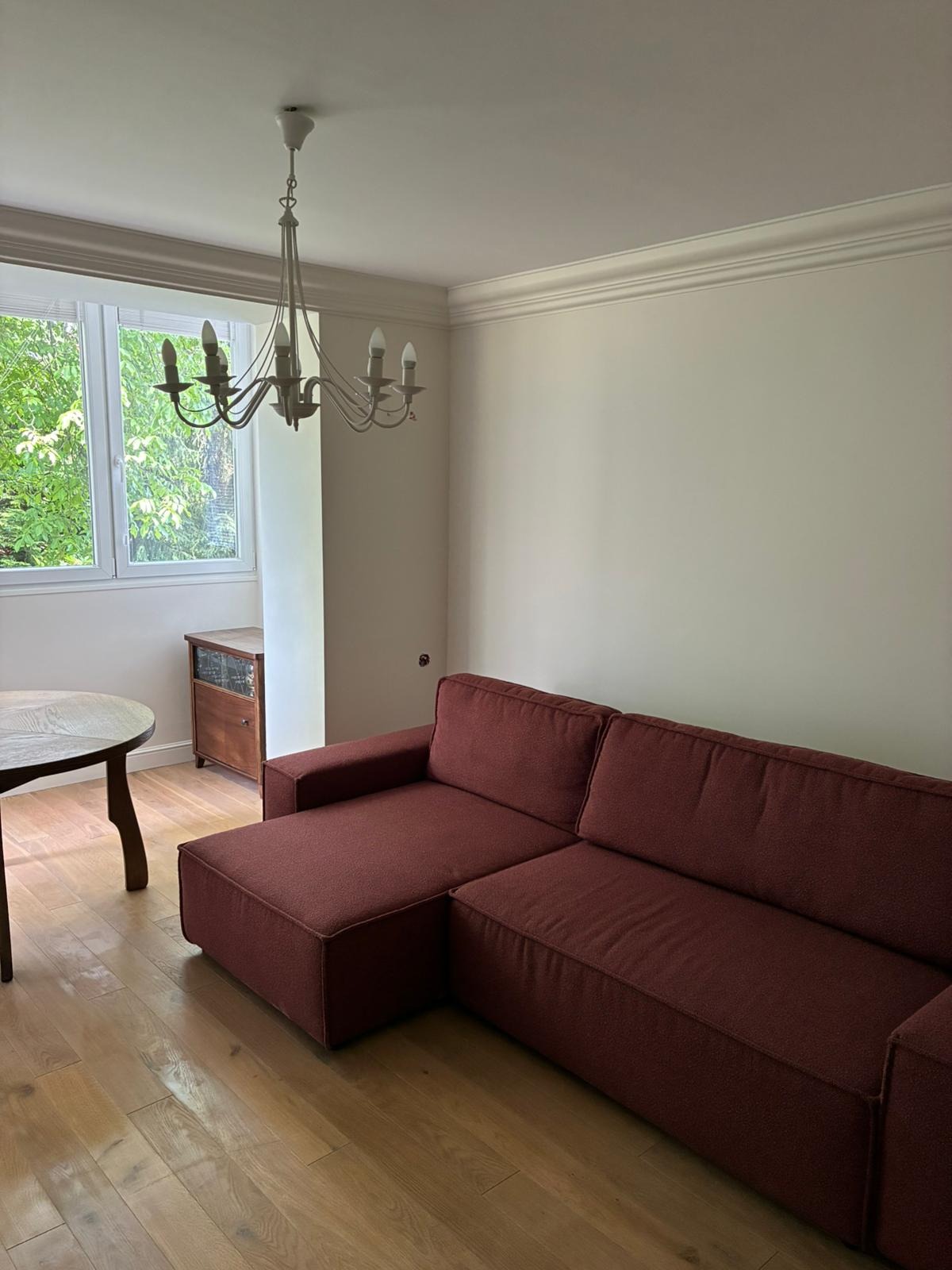 Jasne wnętrze pokoju z drewnianą podłogą, bordową sofą i okrągłym stołem, oświetlone ozdobnym żyrandolem. Ściany pomalowane na jasny odcień.