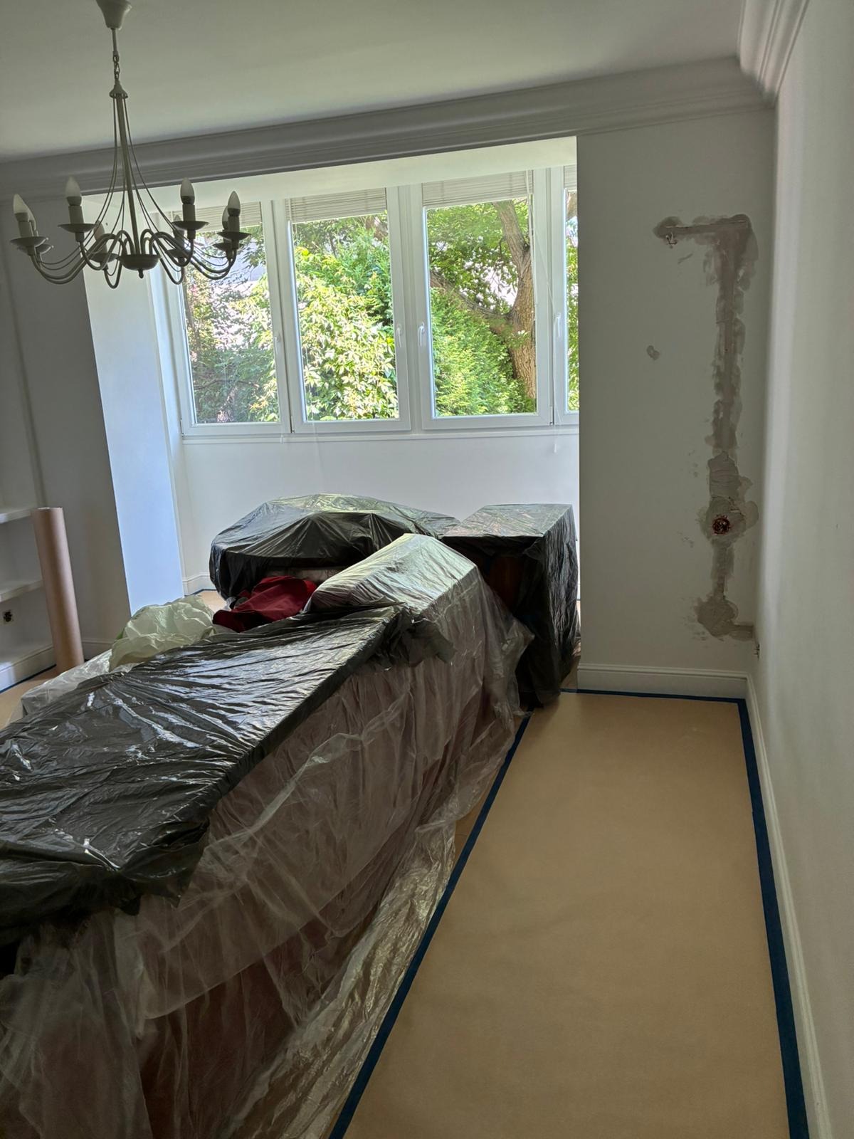 Pokój przed pracami wykończeniowymi: meble zabezpieczone folią, ściana ze śladami szpachlowania, okno z widokiem na zieleń.