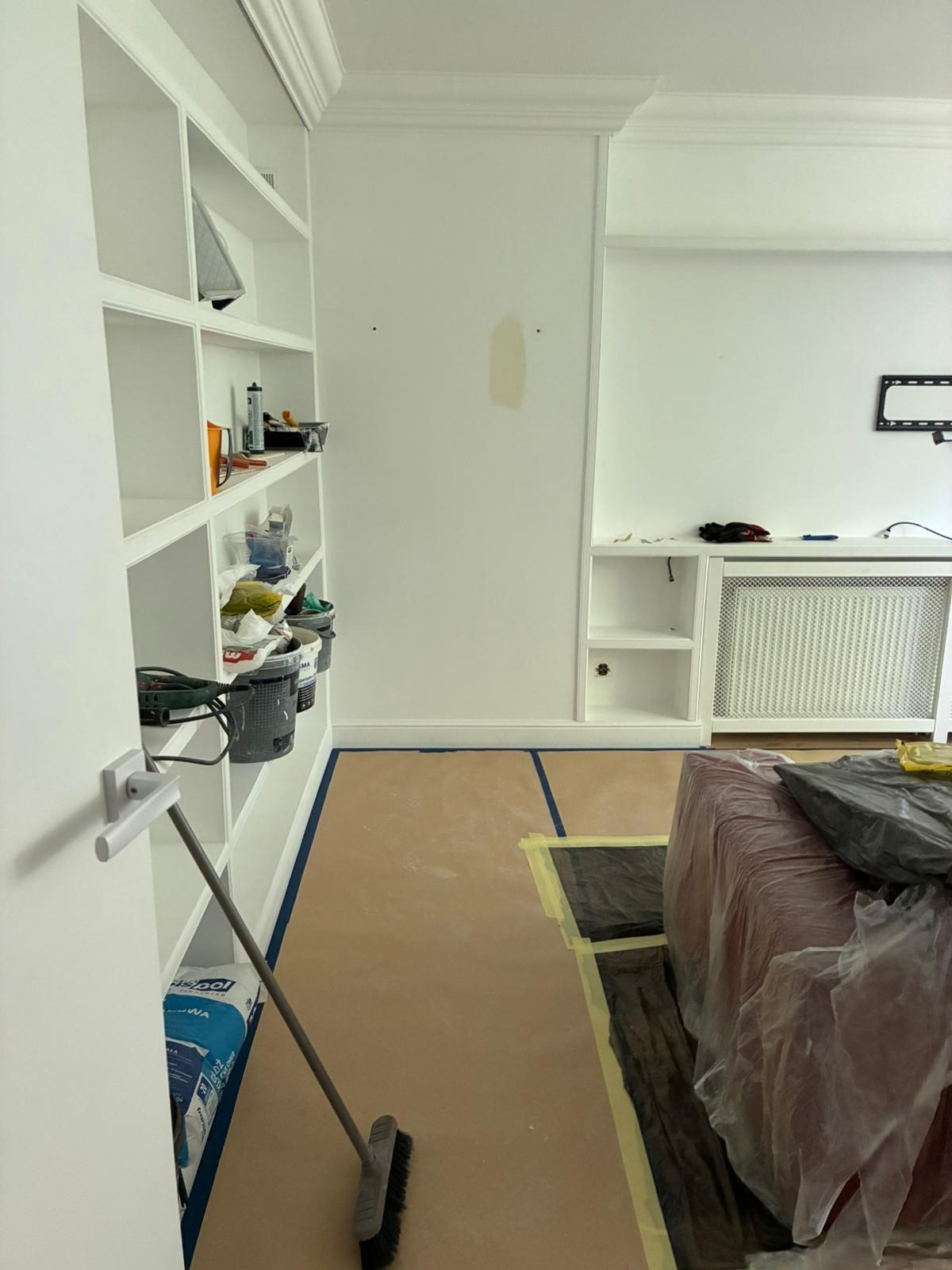 Białe ściany w trakcie malowania, zabezpieczona podłoga, regał z narzędziami i farbami. Widoczne ślady po szpachlowaniu. Przygotowania do malowania mieszkania.