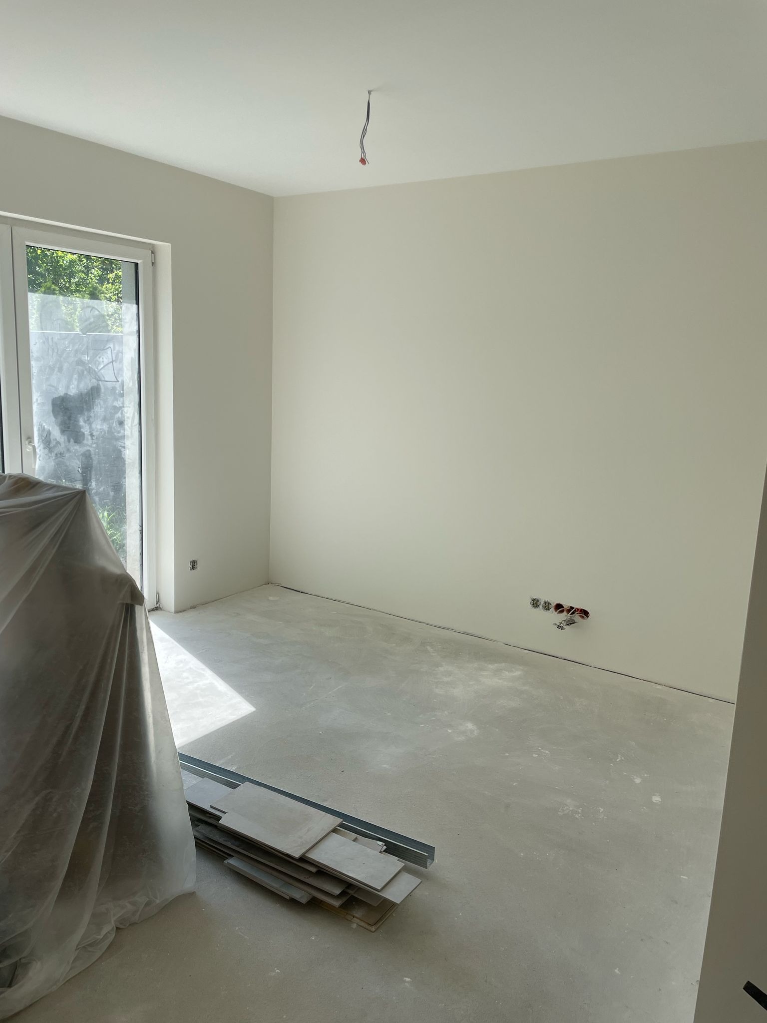 Wnętrze pokoju po szpachlowaniu ścian, gotowe do malowania. Widoczne puszki elektryczne, okno i zakryte meble folią ochronną. Na podłodze leżą płytki.