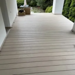 Nowoczesny taras z desek kompozytowych w jasnym odcieniu szarości, widok z poziomu podłogi, z widocznymi elementami konstrukcyjnymi i fragmentem zieleni w tle.