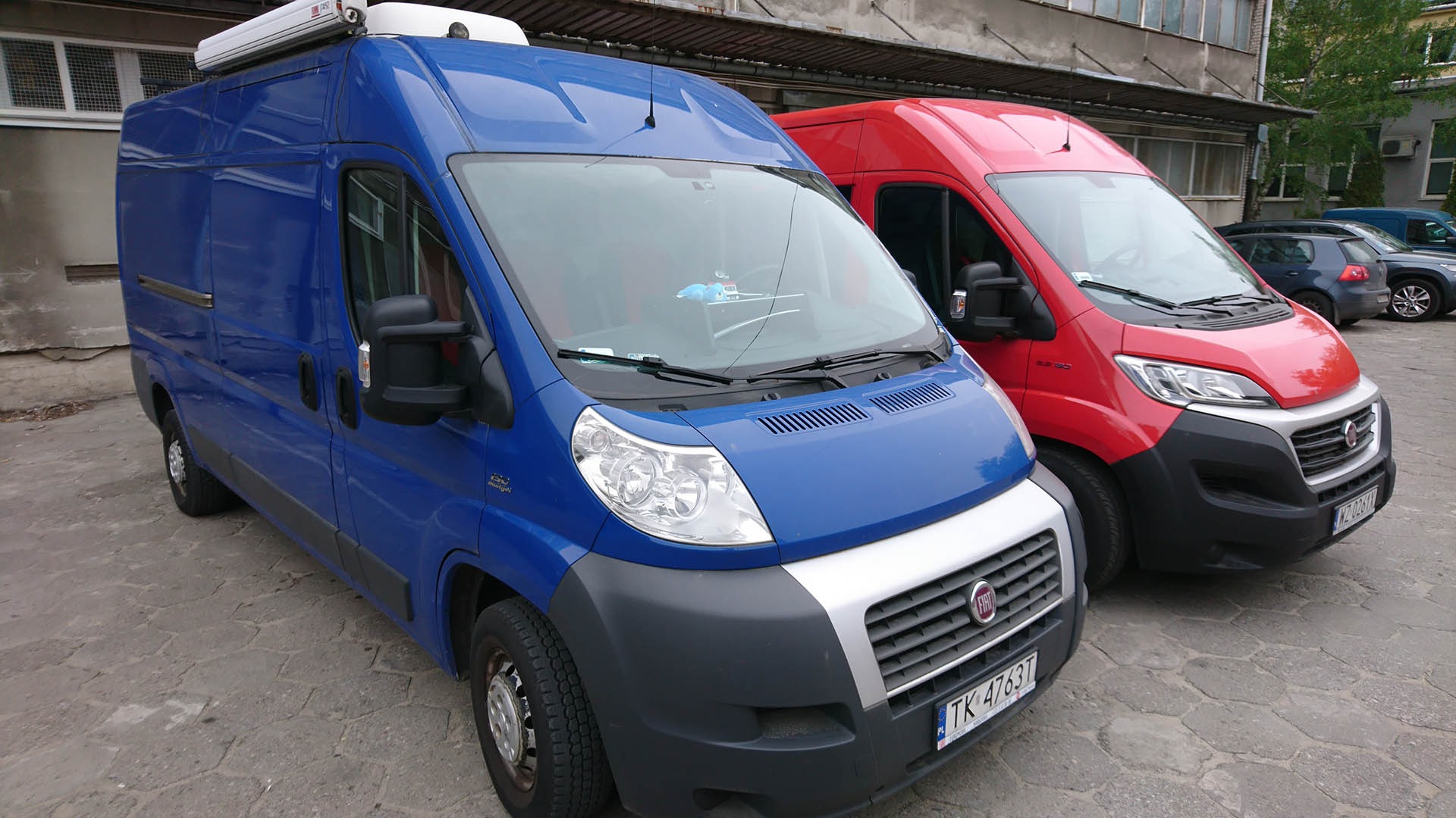 Dwa samochody dostawcze Fiat Ducato: niebieski z agregatem chłodniczym na dachu i czerwony, zaparkowane na zewnątrz budynku.