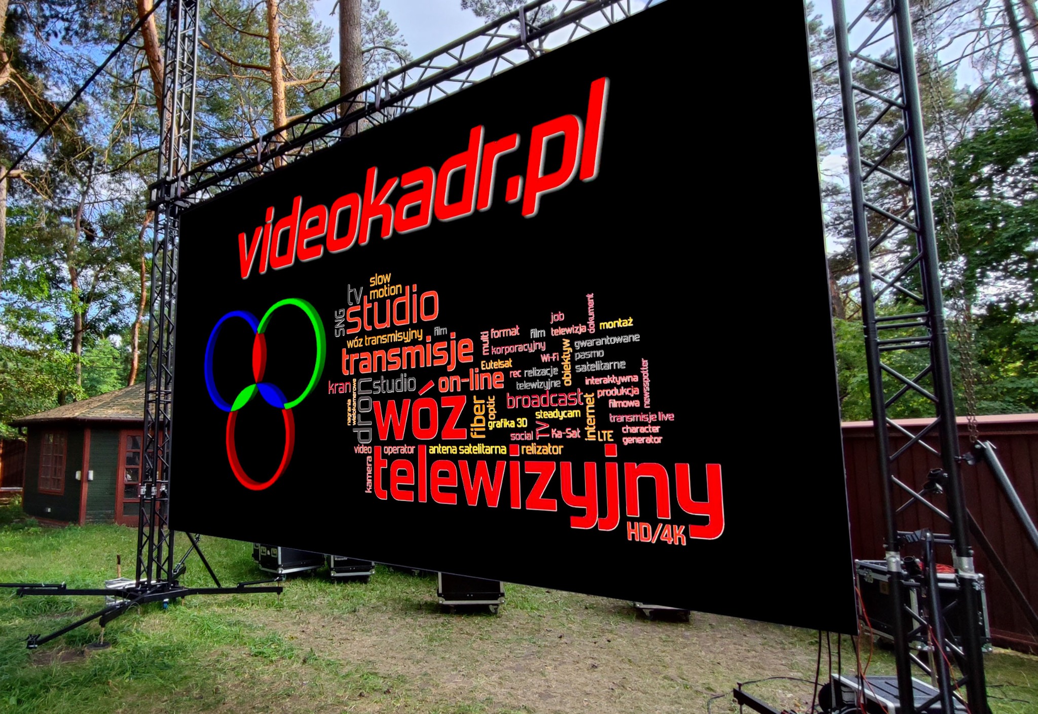 Duży ekran LED na metalowej konstrukcji wyświetla logo i słowa kluczowe związane z produkcją telewizyjną i filmową, ustawiony na trawie w plenerze.