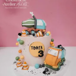 Tort urodzinowy dla Tomka z motywem placu budowy: dwa modele ciężarówek z lukru, stożki drogowe, pastelowe kuleczki, napis 'Tomek 3' na żółtym tle.