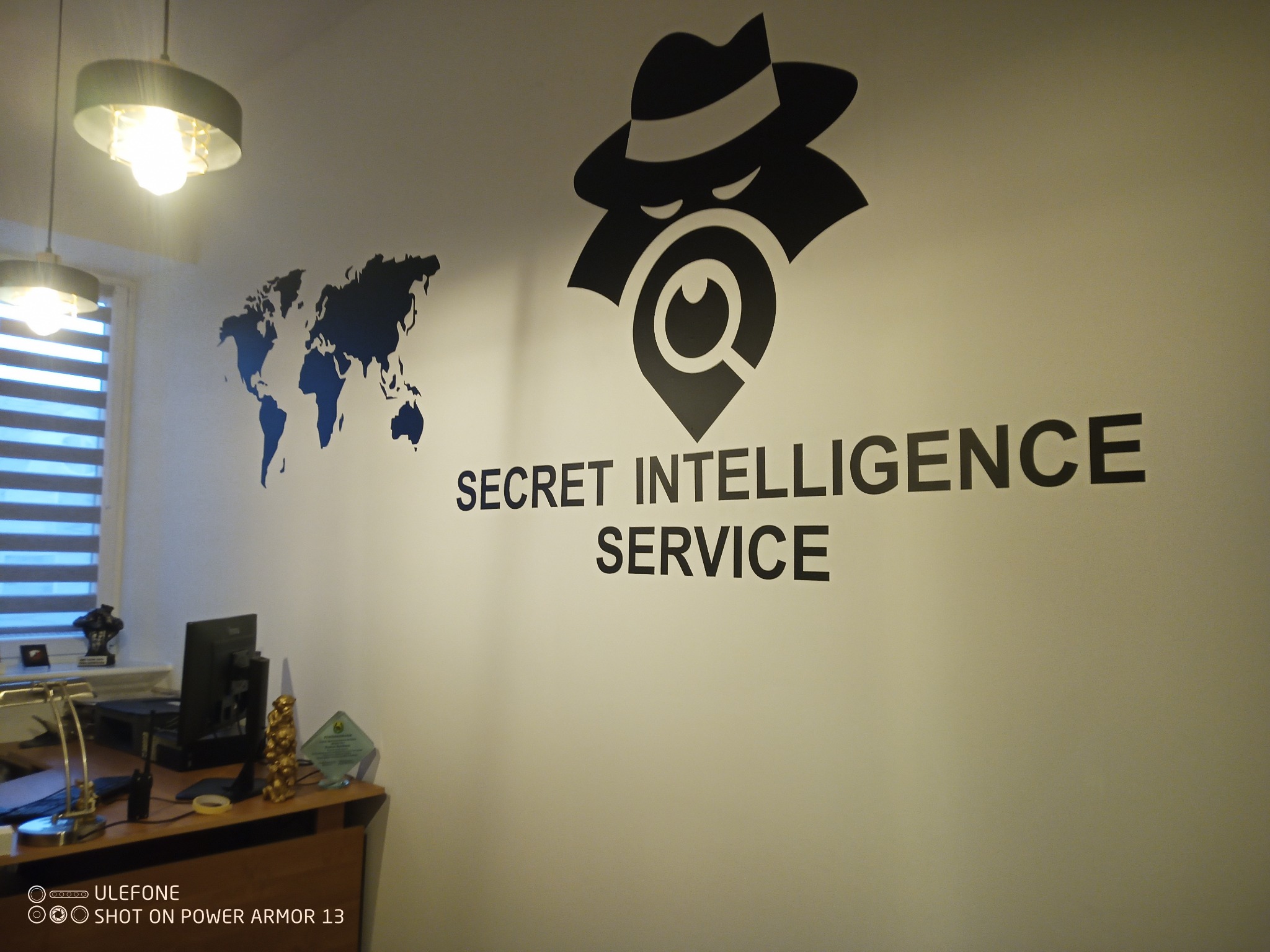 Ściana biura z logo Secret Intelligence Service, mapą świata i biurkiem z komputerem.