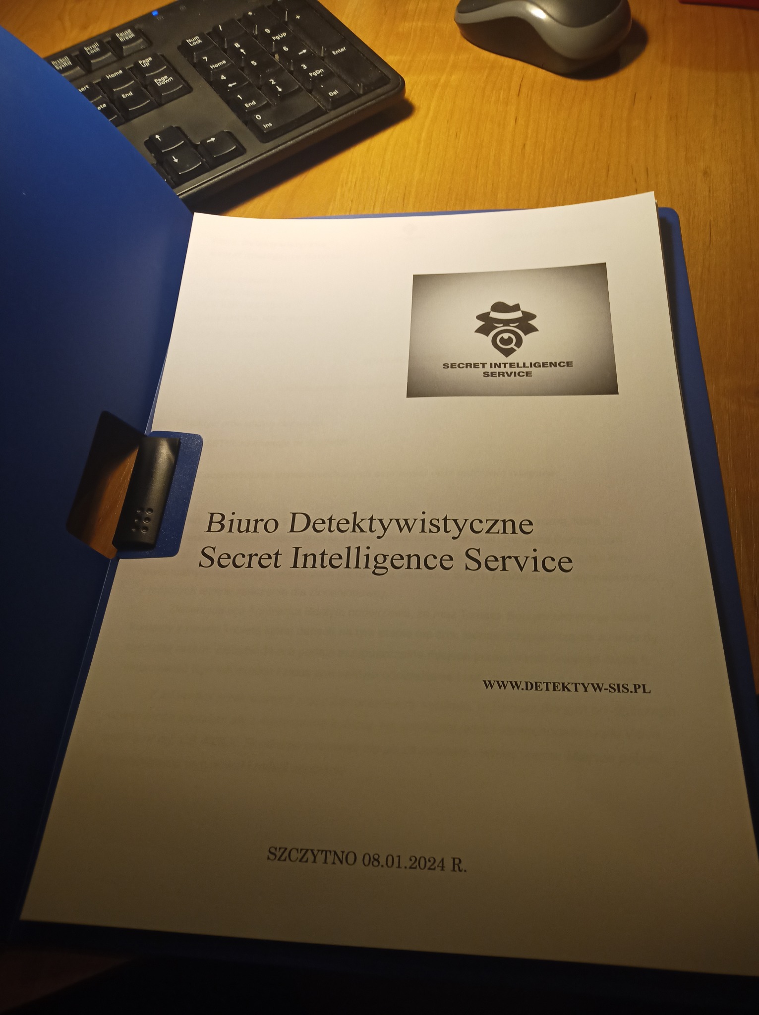 Dokument w niebieskiej teczce z logo i danymi kontaktowymi biura świadczącego usługi wywiadowcze i detektywistyczne, leżący na biurku obok klawiatury i myszki.