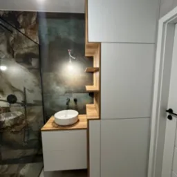 Nowoczesna łazienka z umywalką nablatową, szafką pod umywalką z białymi frontami, drewnianym blatem i regałem z półkami, obok zabudowa meblowa z białymi frontami, widoczny fragment kabiny...