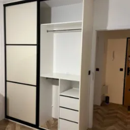 Biała szafa wnękowa z przesuwnymi drzwiami w czarnych ramach, widoczne półki i szuflady, parkiet w jodełkę na podłodze.