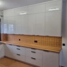 Białe, lakierowane szafki kuchenne z drewnianym blatem i brązowymi, kwadratowymi płytkami na ścianie, oświetlenie sufitowe typu ring.