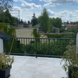 Nowoczesna balustrada metalowa na balkonie z widokiem na zieleń i zabudowania, z donicami z roślinami po bokach, w słoneczny dzień.