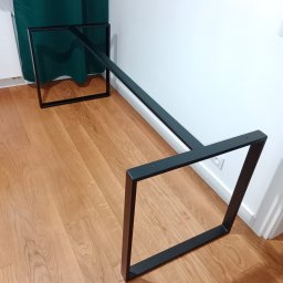 J.Chmielewski Conprometal Balustrady - Metalowa podstawa stołu w kształcie prostokątów połączonych belką, pomalowana na czarno, widoczne otwory montażowe, ułożona na parkiecie w pomieszczeniu.