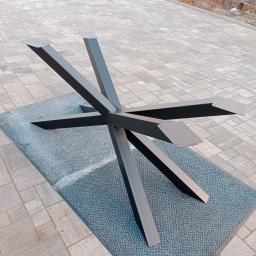 J.Chmielewski Conprometal Balustrady - Metalowa podstawa stołu o nowoczesnym designie z pięcioma nogami, pomalowana na grafitowo, leżąca na szarej wycieraczce, na tle kostki brukowej.