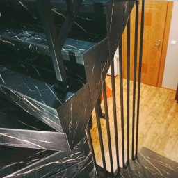 J.Chmielewski Conprometal Balustrady - Schody wewnętrzne z czarnego marmuru z białymi żyłami i metalową balustradą z pionowymi prętami, widok z góry na fragment schodów, w tle drewniane drzwi i podłoga.