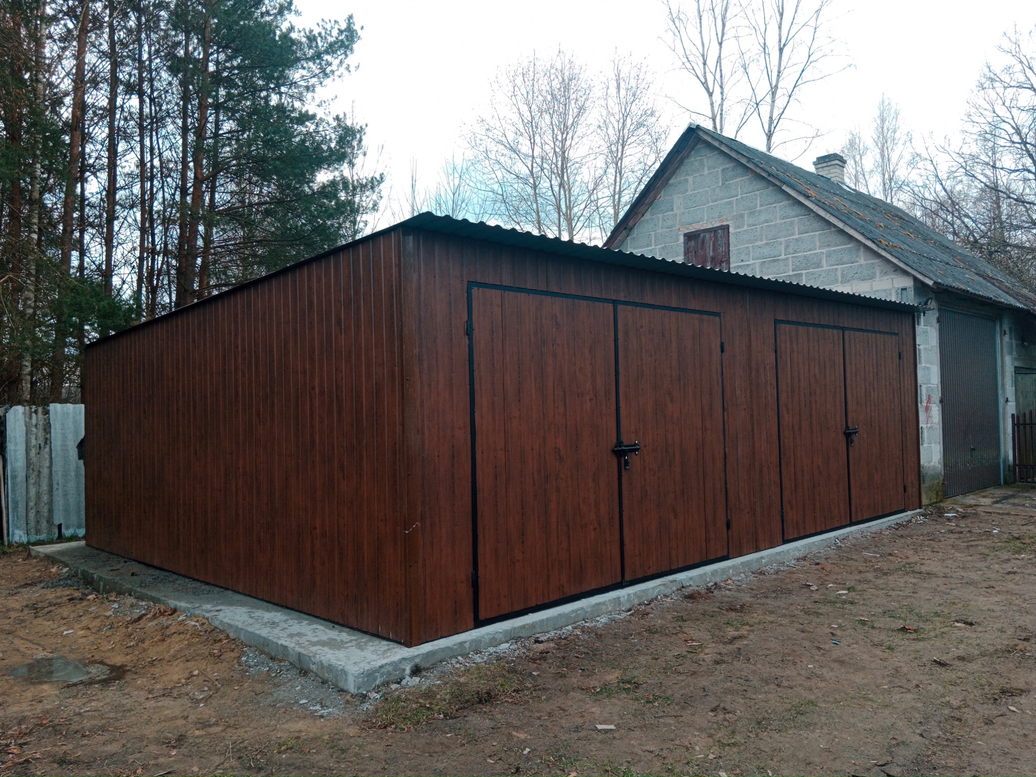 Brązowy garaż blaszany z trzema bramami, stojący na betonowej wylewce, na tle drzew i murowanego budynku.