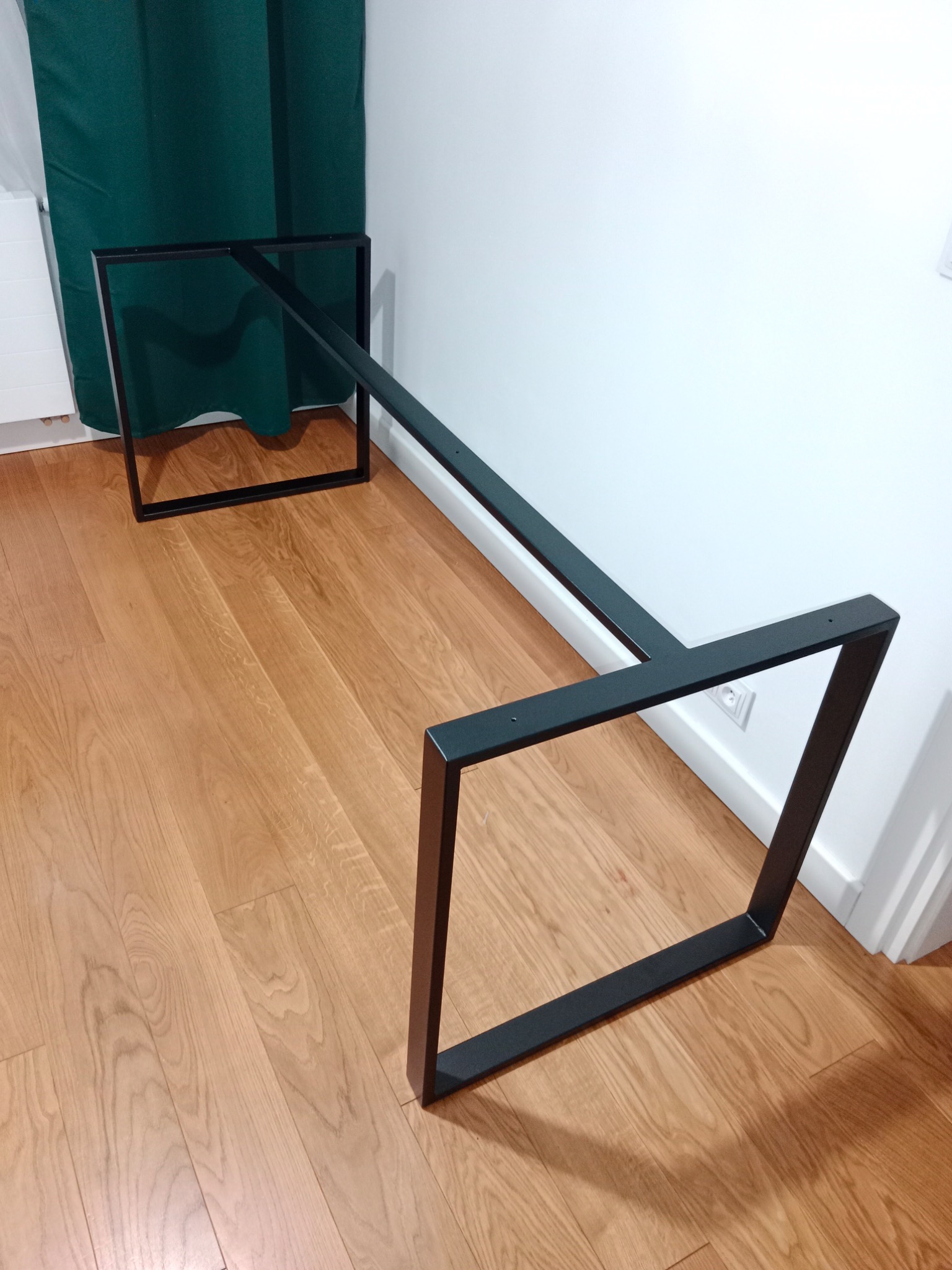 Metalowa podstawa stołu w kształcie prostokątów połączonych belką, pomalowana na czarno, widoczne otwory montażowe, ułożona na parkiecie w pomieszczeniu.