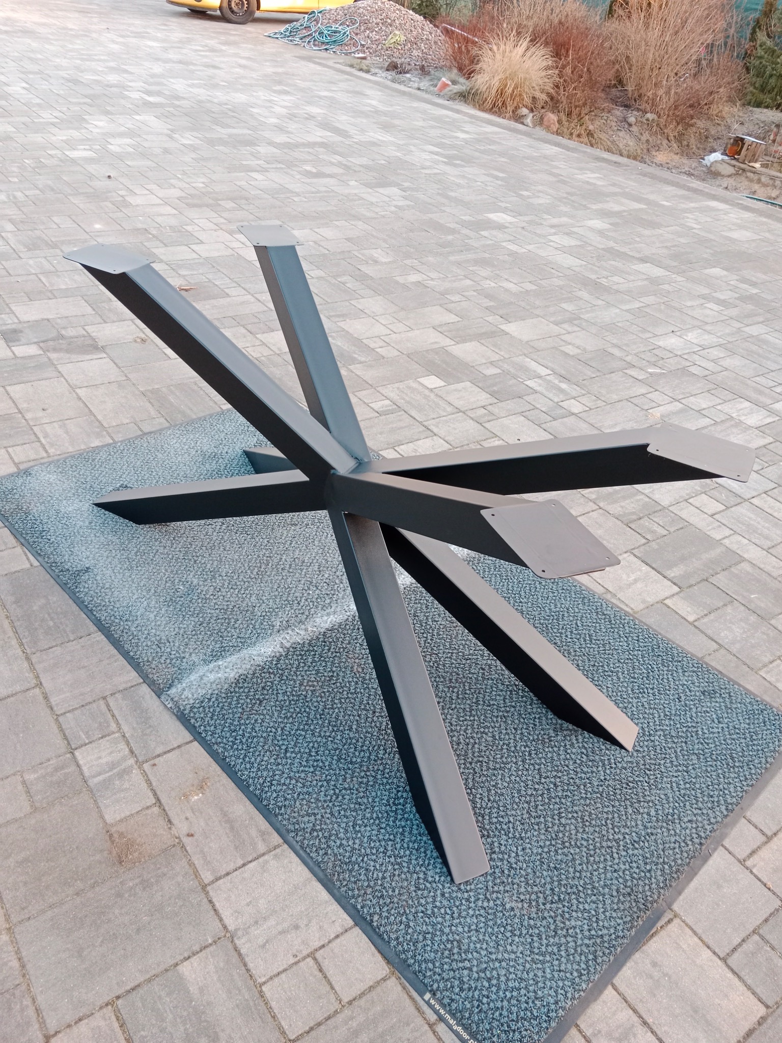 Metalowa podstawa stołu o nowoczesnym designie z pięcioma nogami, pomalowana na grafitowo, leżąca na szarej wycieraczce, na tle kostki brukowej.