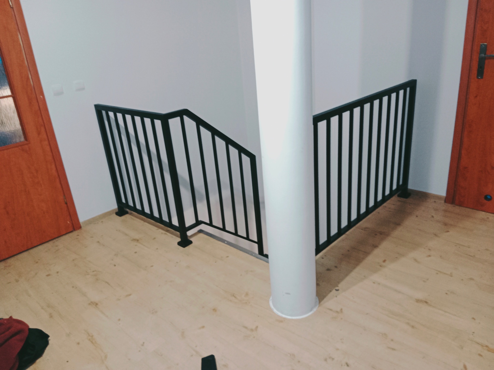 Czarne, metalowe poręcze schodowe z pionowymi szczebelkami, zamontowane w nowoczesnym wnętrzu z drewnianą podłogą i białym słupem nośnym.