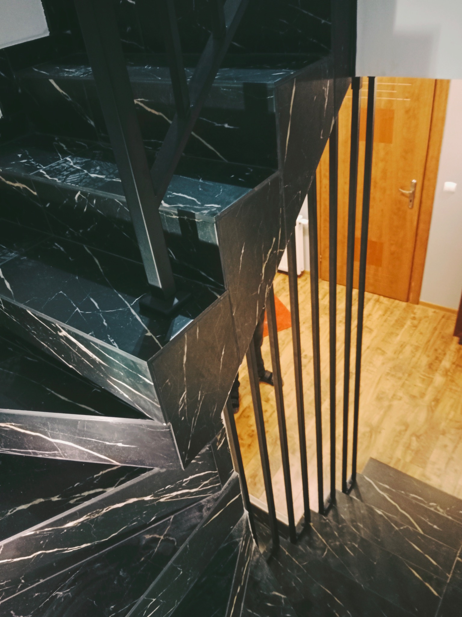 Schody wewnętrzne z czarnego marmuru z białymi żyłami i metalową balustradą z pionowymi prętami, widok z góry na fragment schodów, w tle drewniane drzwi i podłoga.