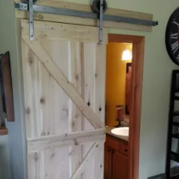 Drewniane drzwi przesuwne w stylu rustykalnym z metalowymi okuciami, częściowo zasłaniające wejście do łazienki z widoczną umywalką i szafką, zamontowane na szynie ponad otworem drzwiowym.