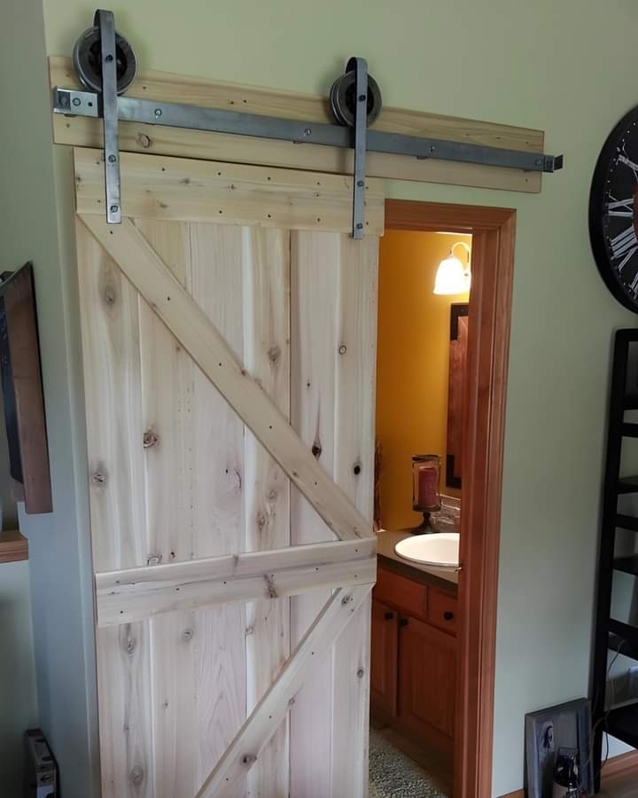 Drewniane drzwi przesuwne w stylu rustykalnym z metalowymi okuciami, częściowo zasłaniające wejście do łazienki z widoczną umywalką i szafką, zamontowane na szynie ponad otworem drzwiowym.