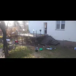 Konstrukcja pod taras z metalowymi słupkami i częściowo zamontowanymi poręczami, widoczna w trakcie budowy przy domu z betonowymi schodami.