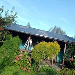 sebastian misiak - Dom z czarnym dachem metalowym, widoczna drabina oparta o ścianę, otoczony zielenią i kwiatami w słoneczny dzień.