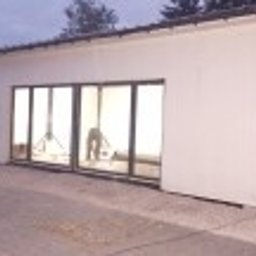 sebastian misiak - Nowoczesny budynek hali z dużymi, przesuwnymi oknami i widocznym wnętrzem w trakcie prac wykończeniowych, oświetlonymi lampami budowlanymi, na zewnątrz szary betonowy plac.