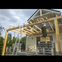 sebastian misiak - Montaż drewnianej konstrukcji z belek na tarasie domu jednorodzinnego, widoczna drabina i mężczyzna pracujący na wysokości.