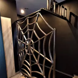 Metalowa barierka o wzorze pajęczyny, zabezpieczająca schody w ciemnym wnętrzu, widoczne niedokończone wykończenie podłogi.