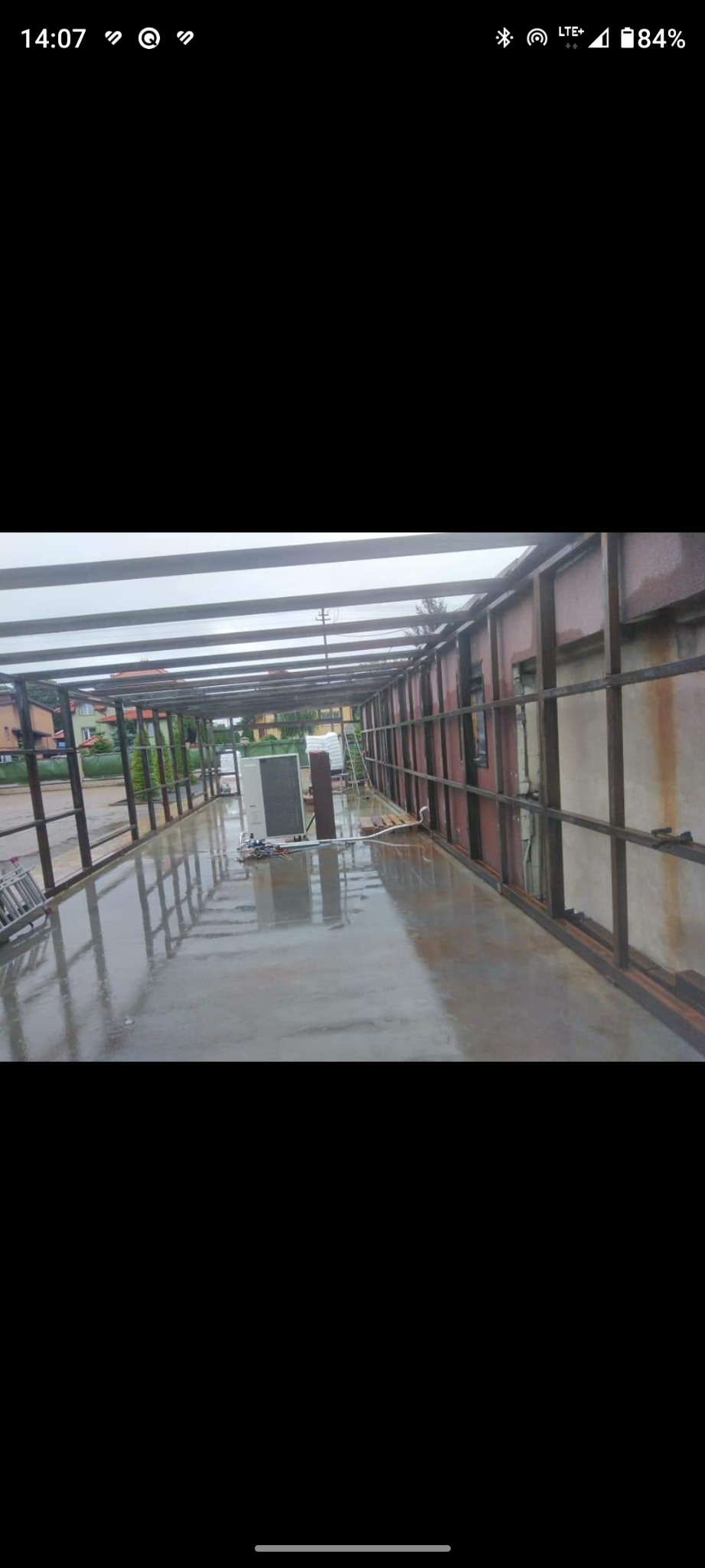 Metalowa konstrukcja zadaszenia w trakcie budowy, widoczne elementy nośne i poprzeczne, mokra betonowa posadzka, klimatyzator na zewnątrz budynku.