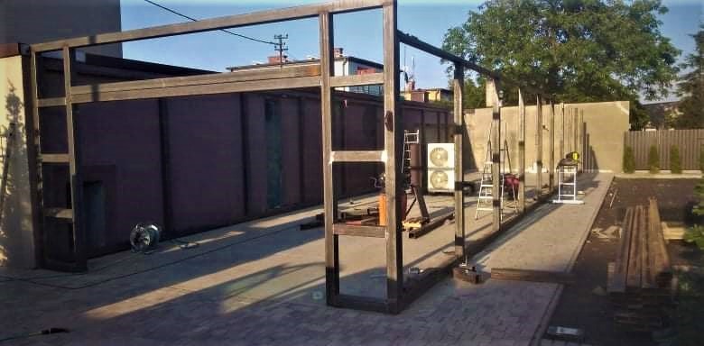 Konstrukcja metalowa w trakcie budowy, widoczne elementy szkieletu i drabiny, tło z budynkiem i zielenią, plac budowy z narzędziami.