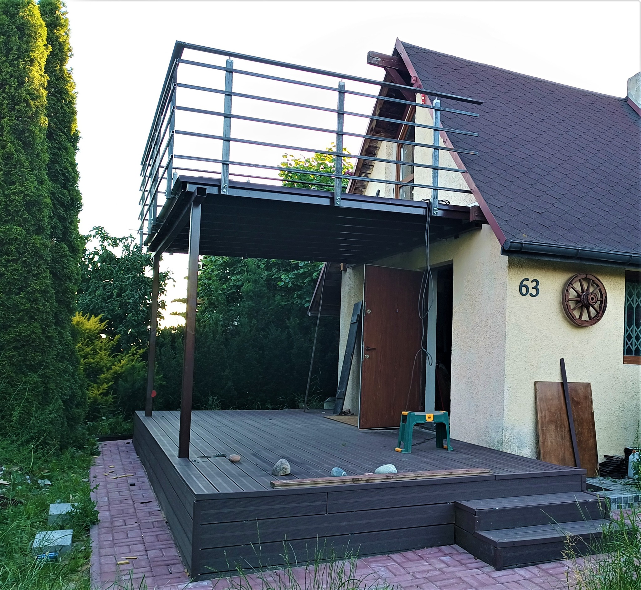 Metalowa konstrukcja balkonu z poręczą, połączona z budynkiem, z drewnianym tarasem na dole, widok z poziomu ogrodu.