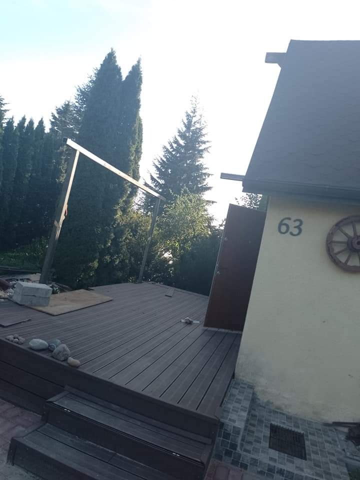 W budowie drewniany taras z kompozytu przy domu z numerem 63, widoczne elementy konstrukcji pergoli, kamienie dekoracyjne i drzewa w tle.