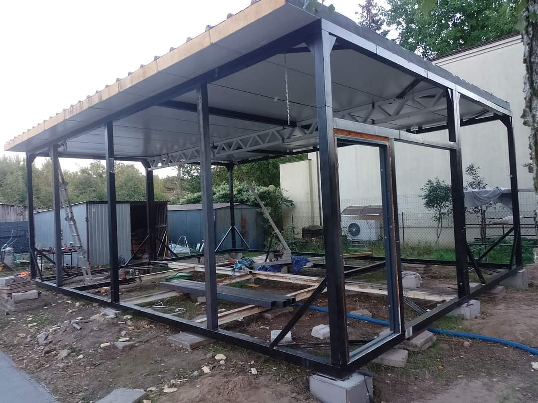 Konstrukcja z czarnych profili stalowych z zadaszeniem w trakcie budowy na działce, widoczne bloczki betonowe i elementy rusztowania.