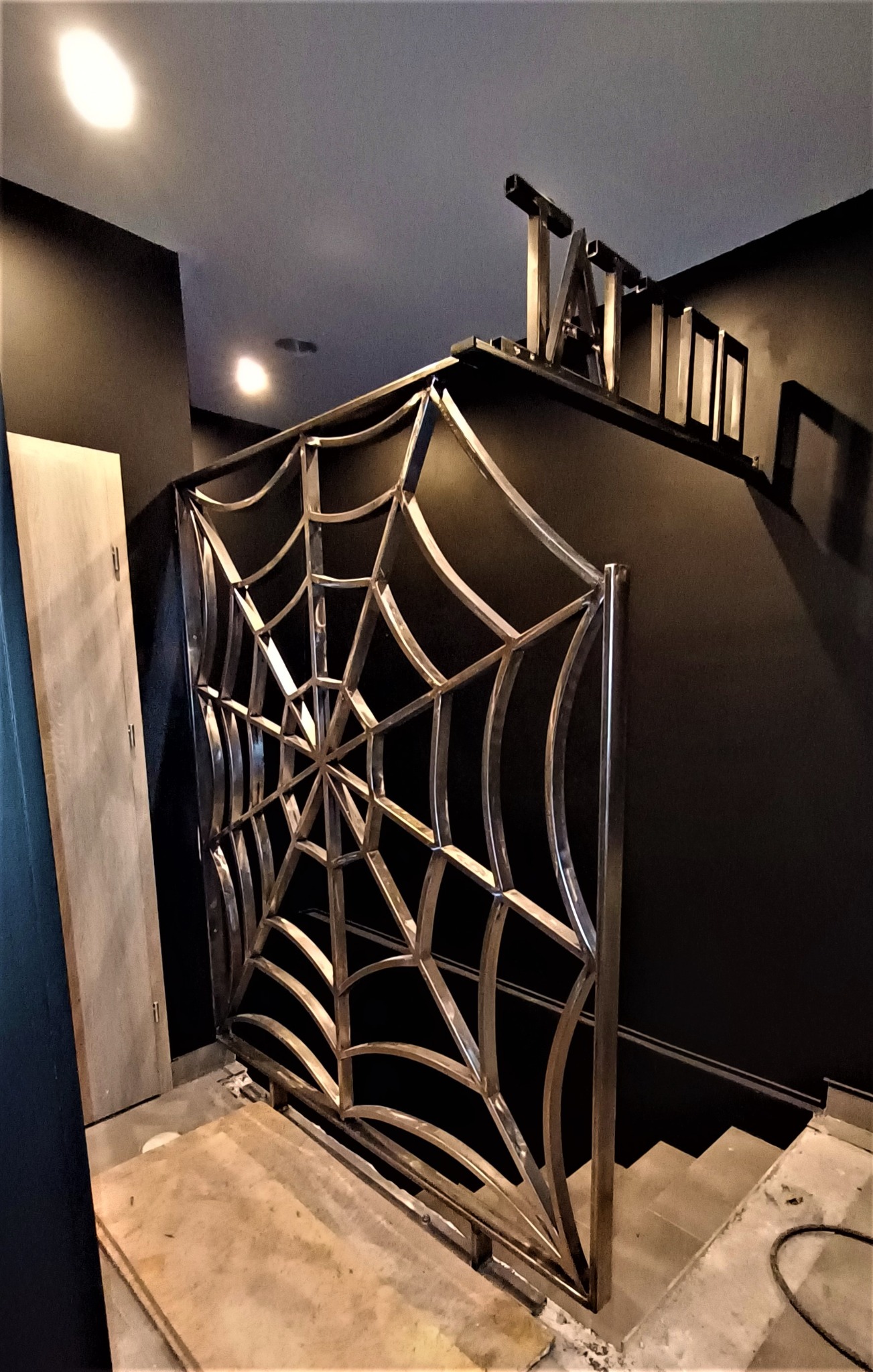 Metalowa barierka o wzorze pajęczyny, zabezpieczająca schody w ciemnym wnętrzu, widoczne niedokończone wykończenie podłogi.