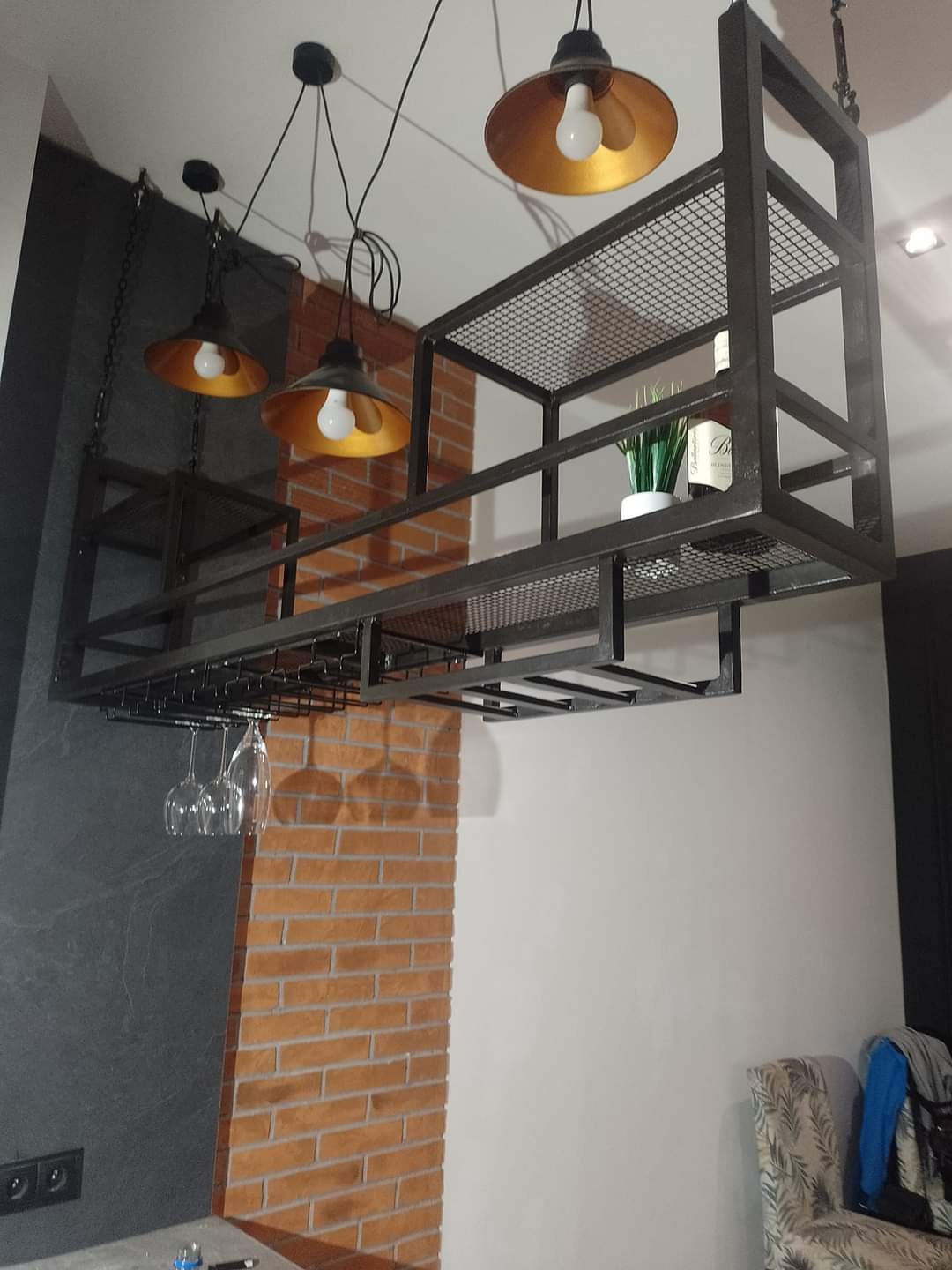 Metalowa konstrukcja podwieszana do sufitu z miejscem na butelki, rośliny i wieszakiem na kieliszki, oświetlona trzema lampami z metalowymi kloszami, na tle ściany z cegły i szarego kamienia.