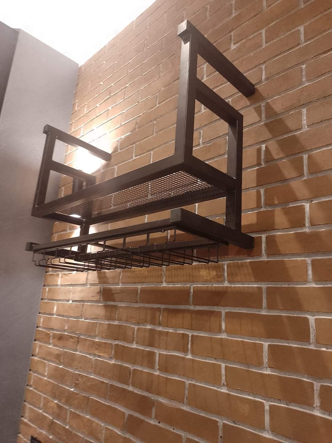 Metalowa konstrukcja do przechowywania kieliszków i butelek, zamontowana na ceglanej ścianie z widocznym oświetleniem punktowym.