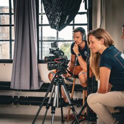 RAFAELLO FILM RAFAŁ MAĆKOWIAK - Ekipa filmowa podczas pracy: mężczyzna w słuchawkach siedzi na stołku za kamerą na statywie, obok dwie kobiety obserwują nagrywany obraz. W tle okno z widokiem na budynek.