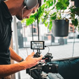 RAFAELLO FILM RAFAŁ MAĆKOWIAK - Mężczyzna w słuchawkach obsługuje profesjonalną kamerę Sony z podglądem na monitorze, w tle widoczne rośliny doniczkowe i okno.