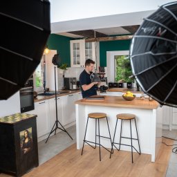 RAFAELLO FILM RAFAŁ MAĆKOWIAK - Mężczyzna w kuchni obsługuje profesjonalną kamerę filmową, otoczony lampami softbox, na blacie leży laptop, w tle białe szafki i zielona ściana.