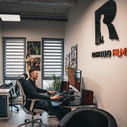 RAFAELLO FILM RAFAŁ MAĆKOWIAK - Mężczyzna w czarnej koszuli siedzi przy biurku z kamerą Sony i monitorem, edytując film. Na ścianie logo firmy 'Raffello Film' w czerni i pomarańczy. W tle okna z roletami i zdjęcia z planu filmowego.