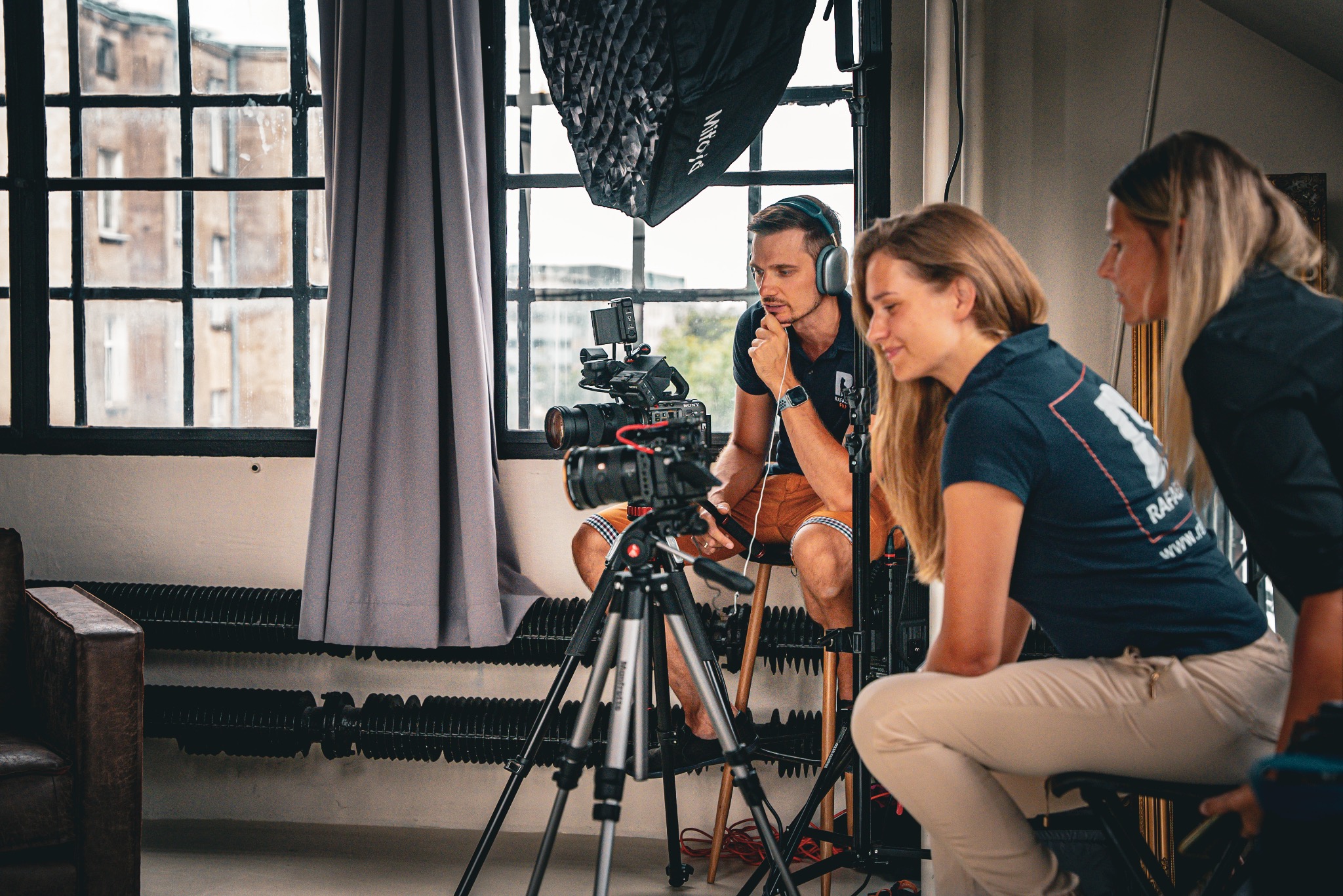 Ekipa filmowa podczas pracy: mężczyzna w słuchawkach siedzi na stołku za kamerą na statywie, obok dwie kobiety obserwują nagrywany obraz. W tle okno z widokiem na budynek.