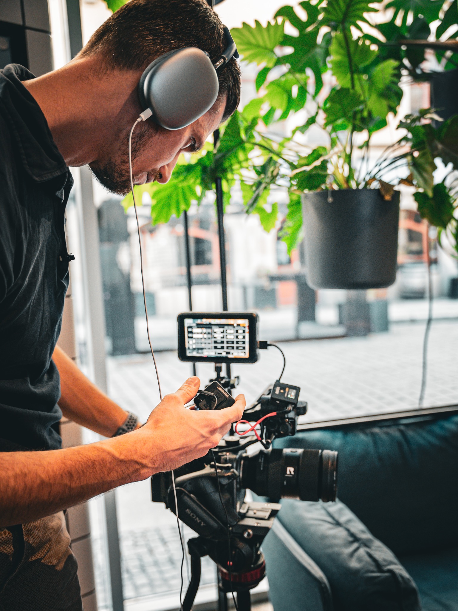 Mężczyzna w słuchawkach obsługuje profesjonalną kamerę Sony z podglądem na monitorze, w tle widoczne rośliny doniczkowe i okno.