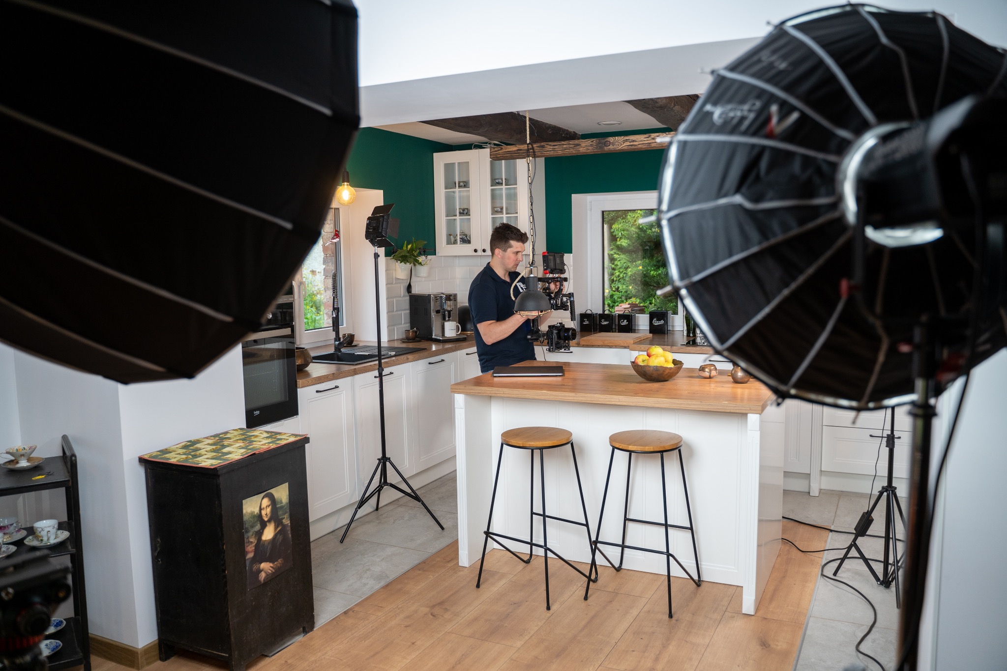 Mężczyzna w kuchni obsługuje profesjonalną kamerę filmową, otoczony lampami softbox, na blacie leży laptop, w tle białe szafki i zielona ściana.