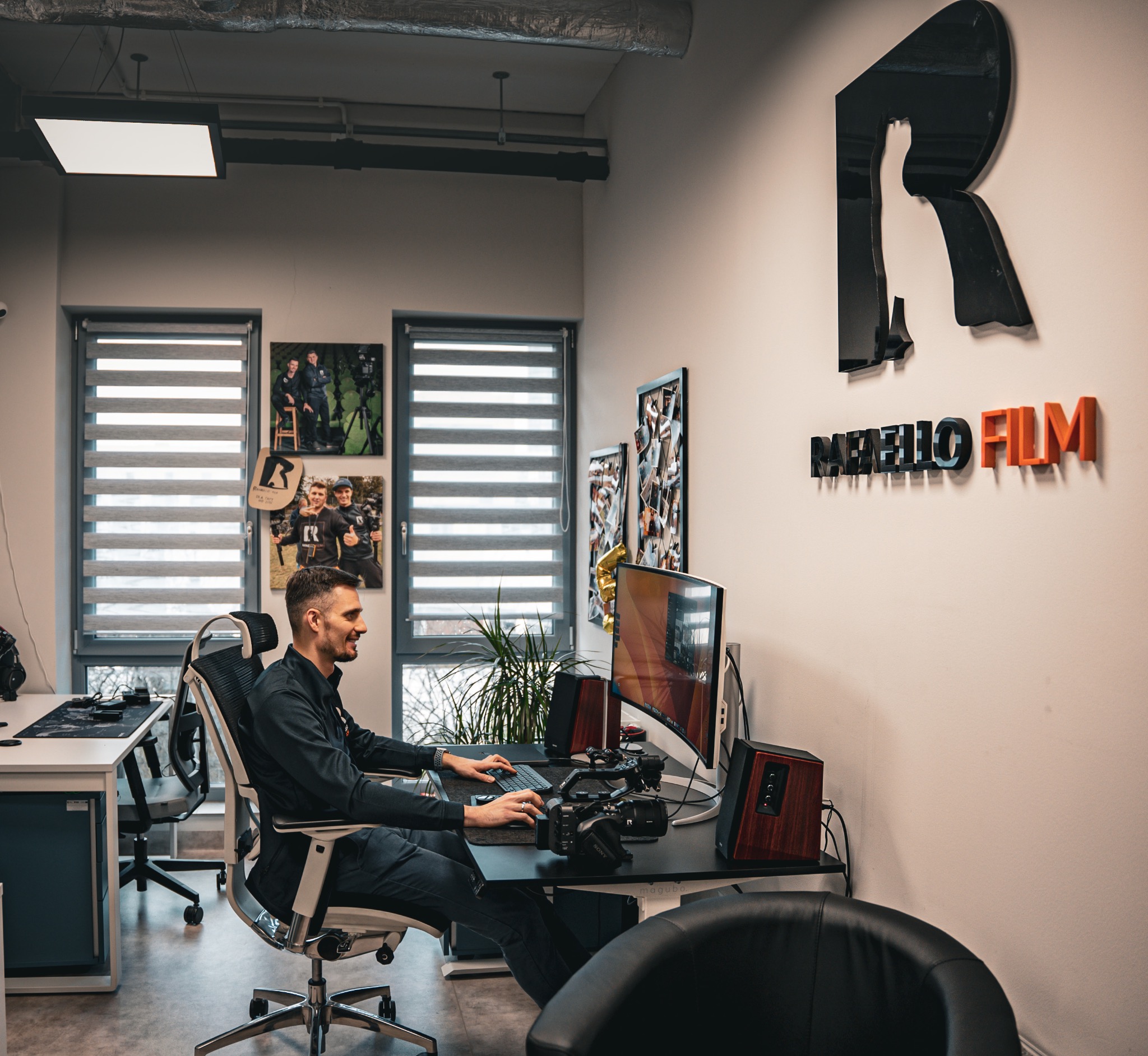 Mężczyzna w czarnej koszuli siedzi przy biurku z kamerą Sony i monitorem, edytując film. Na ścianie logo firmy 'Raffello Film' w czerni i pomarańczy. W tle okna z roletami i zdjęcia z planu filmowego.