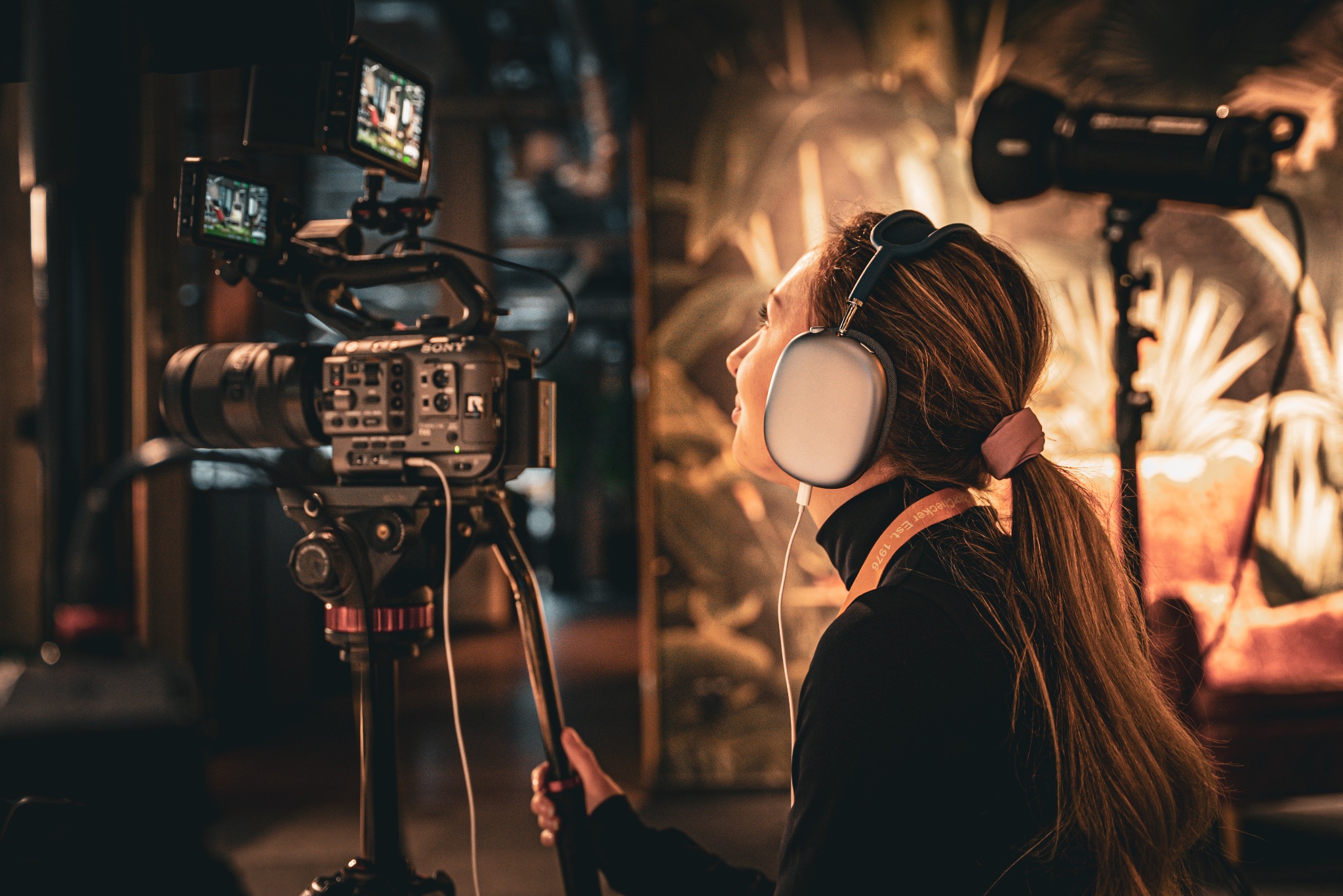 Kobieta w słuchawkach obsługuje profesjonalną kamerę Sony na statywie z dwoma monitorami, oświetloną lampą studyjną w tle.