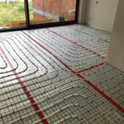 Rozłożone rury ogrzewania podłogowego na siatce montażowej, widoczne czerwone taśmy mocujące, w tle surowy mur z cegieł i okno z widokiem na zieleń.