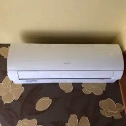 Biała klimatyzacja marki Gree zamontowana na ścianie z brązową tapetą w kwiaty, widoczna czerwona dioda sygnalizacyjna.