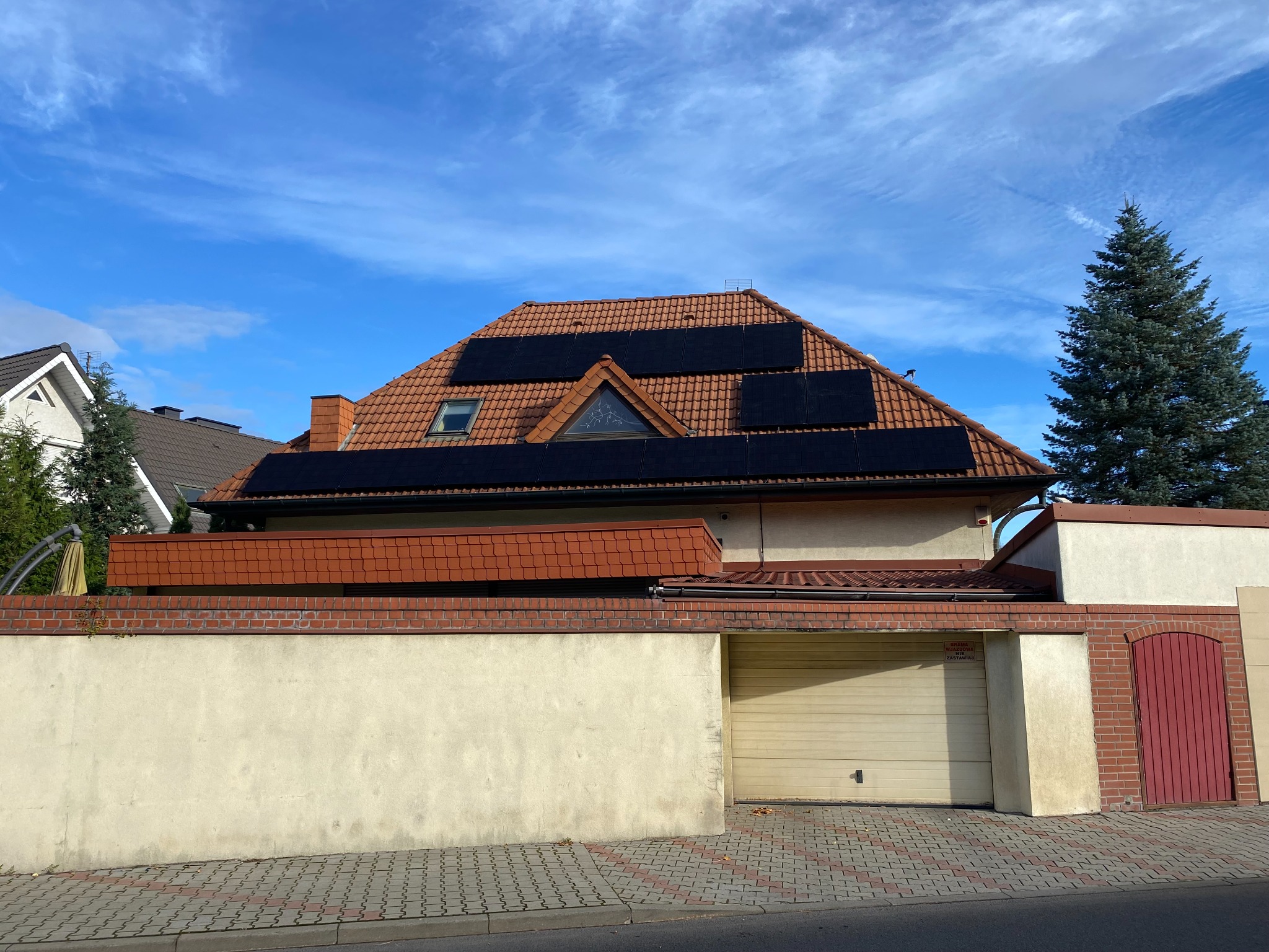 Dom jednorodzinny z panelami fotowoltaicznymi na dachu z czerwonej dachówki, widok z ulicy w słoneczny dzień.