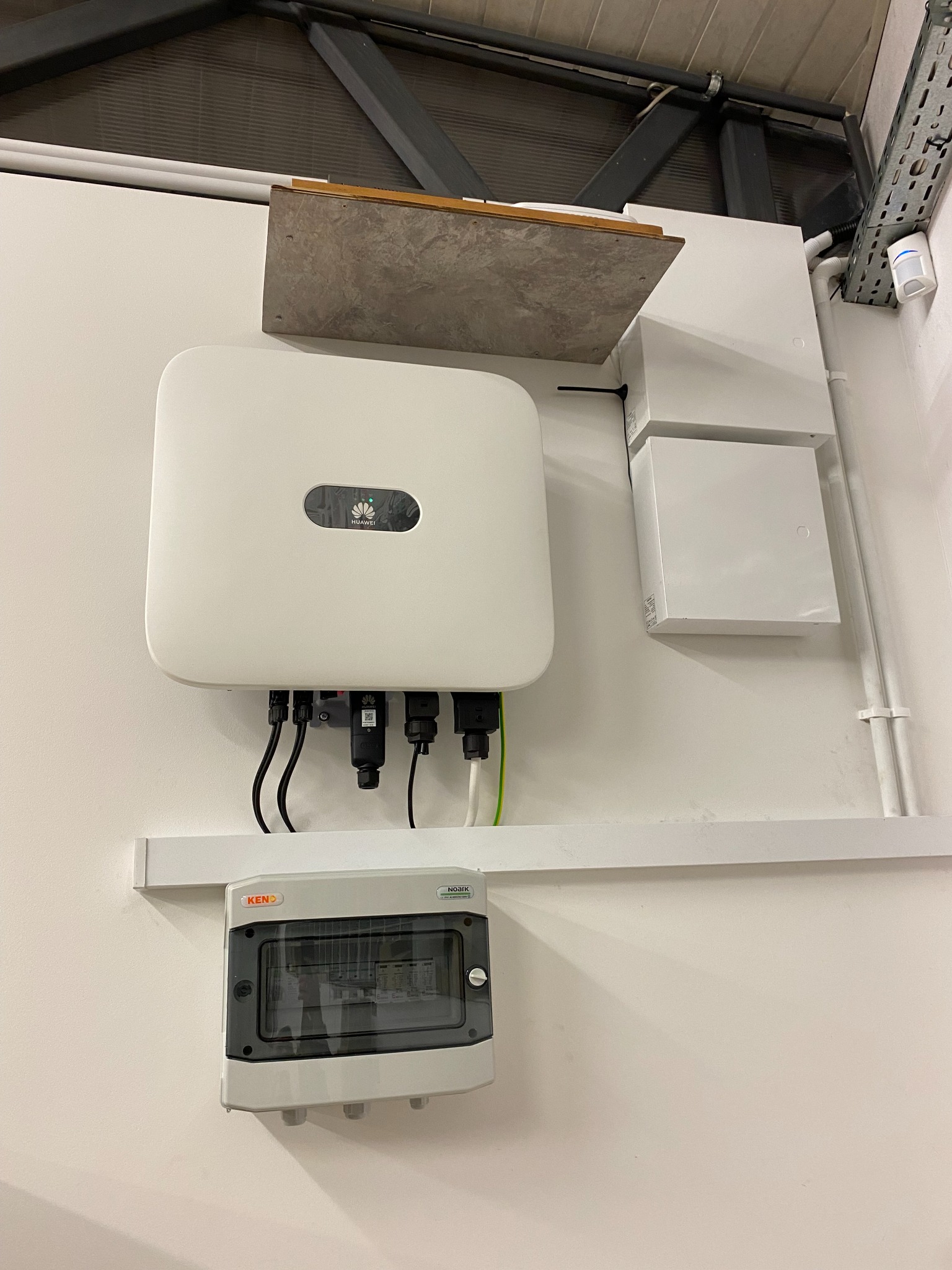 Biały inwerter fotowoltaiczny Huawei zamontowany na ścianie, widoczne kable, puszka instalacyjna KENO pod spodem, białe rurki instalacyjne i puszki po prawej stronie, instalacja wewnątrz budynku.